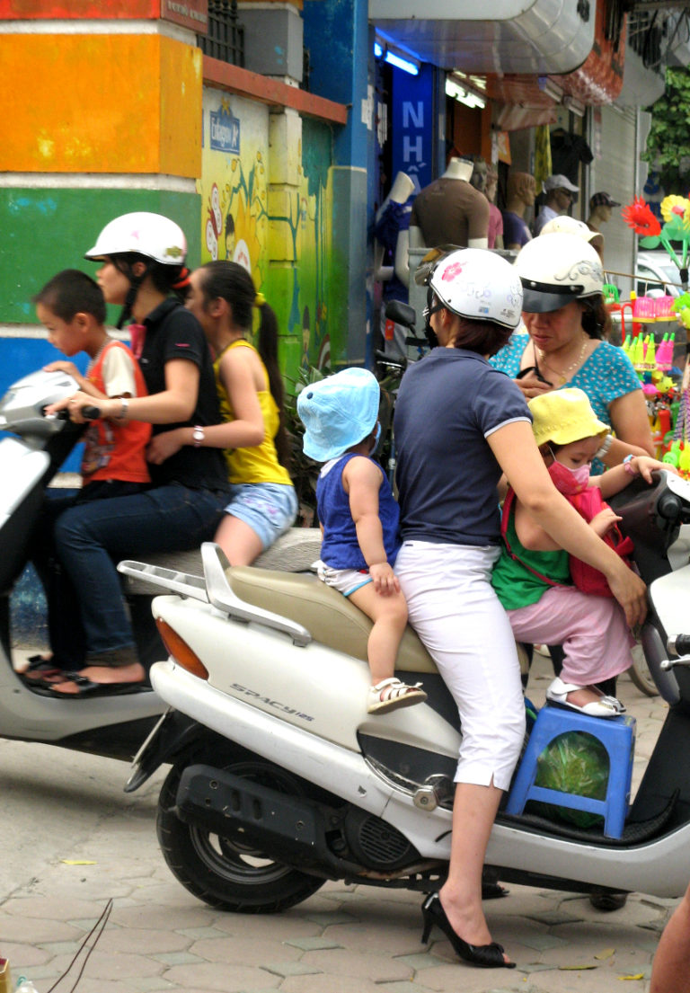 Deux jeunes mères en scooter avec leurs enfants dans une rue vietnamienne, les enfants à l'avant-plan sont très jeunes. Les adultes portent des casques de vélo mais pas les enfants.