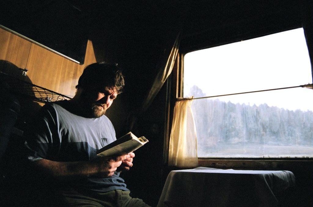 Un homme lit un livre de poche dans un compartiment d'un vieux train, on voit une forêt à l'extérieur