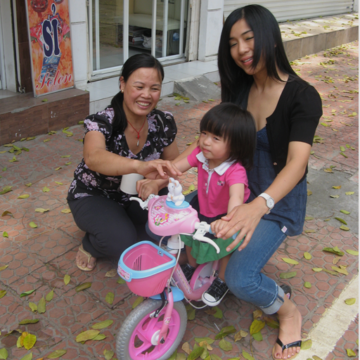 Gouter dans la rue: une enfant de 2 ou 3 ans sur un tricycle rose avec deux femmes souriantes