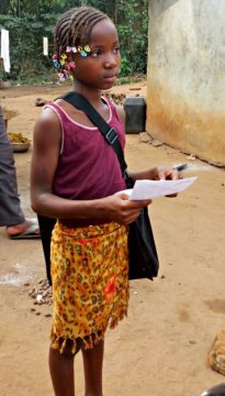 Une fillette (joliment coiffée) attend dans la rue d'un village, elle porte un sacoche et tient une feuille de papier à la main