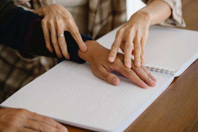 Sur une page de texte en Braille, on voit la main d'un malboyant guidées par les deux mains d'une autre personne d'une certaine age - on ne voit pas leur visage