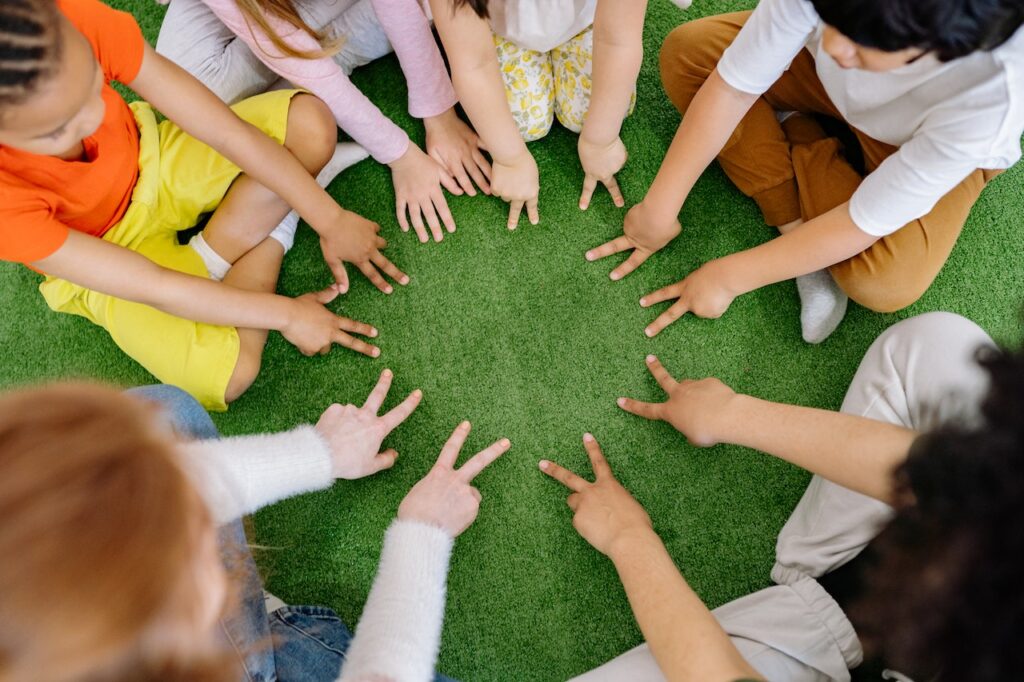 Des mains d'enfants forment un cercle, chacun tend 2 doigts de chaque main vers le centre, sauf une personne dont on voit les 5 doignts de chaque main. On ne voit pas leurs visages.