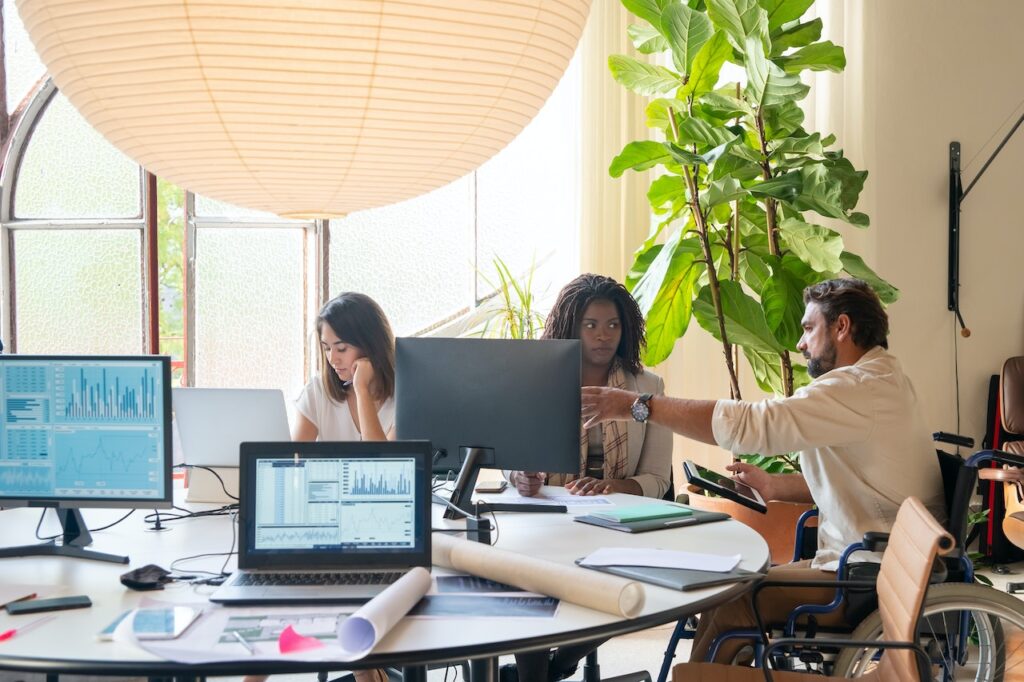 Autour d'une table où l'on voit des écrans d'ordinateur et documents; deux femmes et un homme discutent d'un projet de travail L'homme est en chaise roulante.