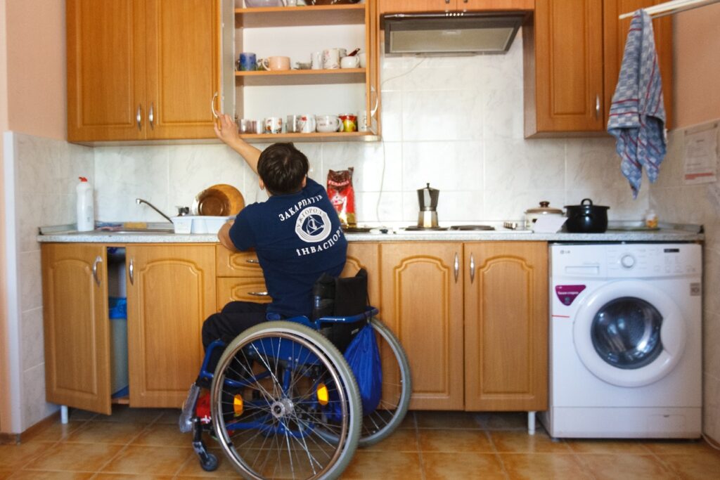 On jeune homme en chaise roulante dans la cuisine chez lui où il tend le bras pour ouvrir une armoire haute. Tout doit être à sa portée.