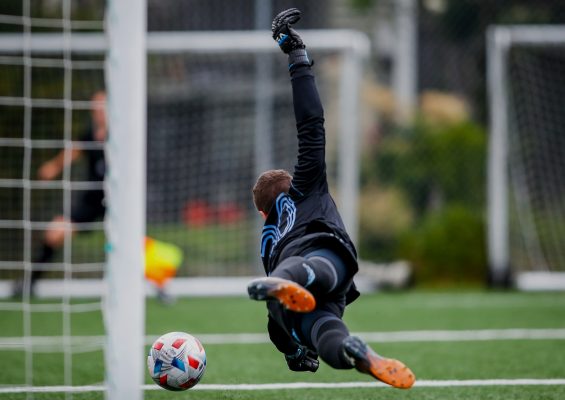 Terrain de football où un jeune gardien de but se jette dans l'air pour arrêter un but, en avant-plan le ballon qui frole le poteau et les pieds du gardien