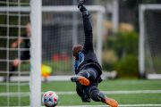 Terrain de football où un jeune gardien de but se jette dans l'air pour arrêter un but, en avant-plan le ballon qui frole le poteau et les pieds du gardien